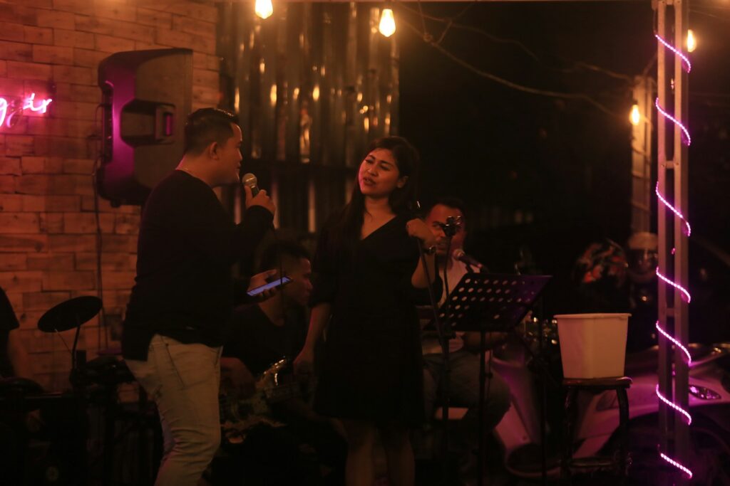 A male singer performs on stage with a microphone, engaging with a female vocalist. The setting features warm lighting and musicians in the background, creating a lively atmosphere.