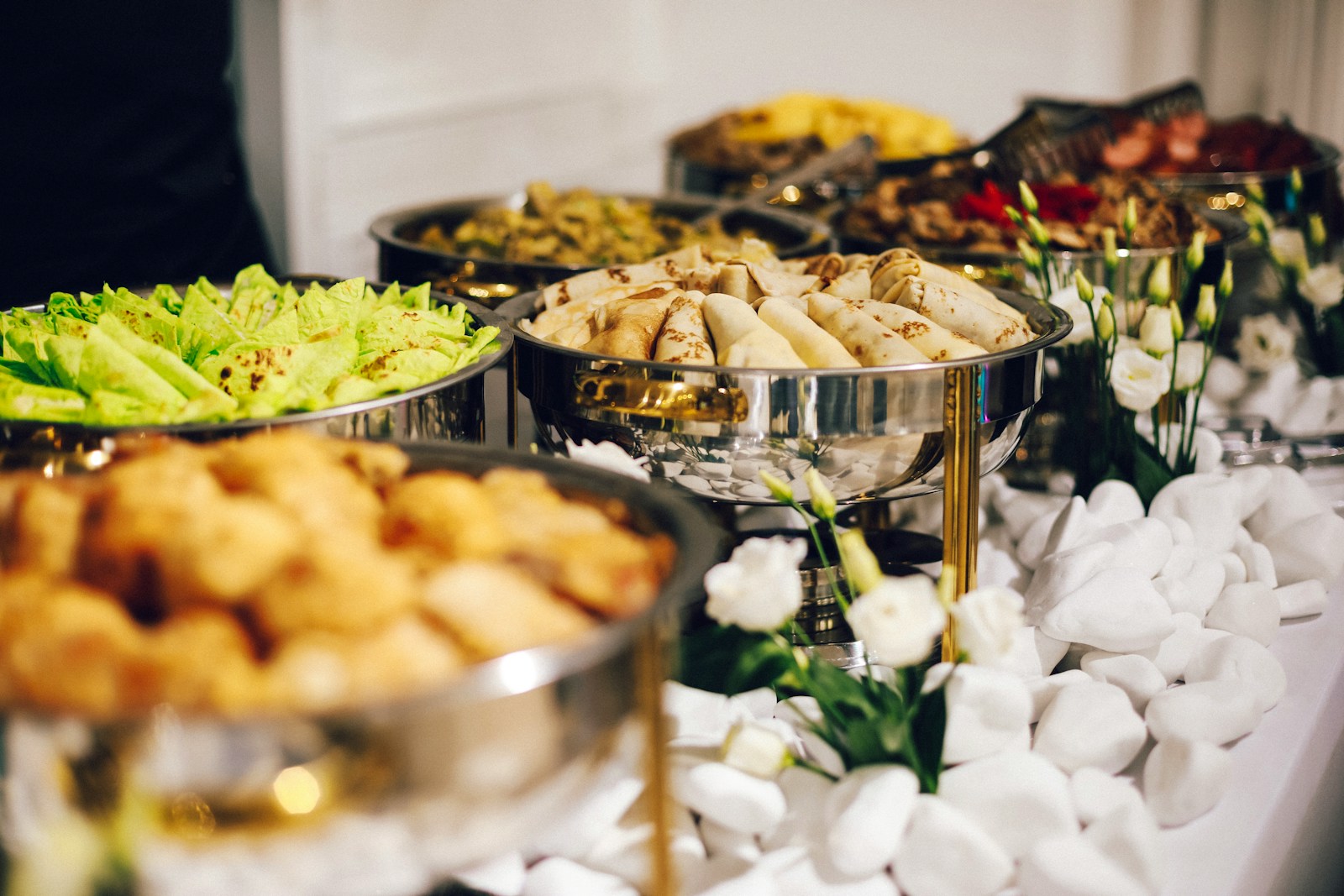 A buffet featuring a variety of dishes in stainless steel serving platters, including leafy greens, wraps, and bread, surrounded by white decorative stones and flowers.