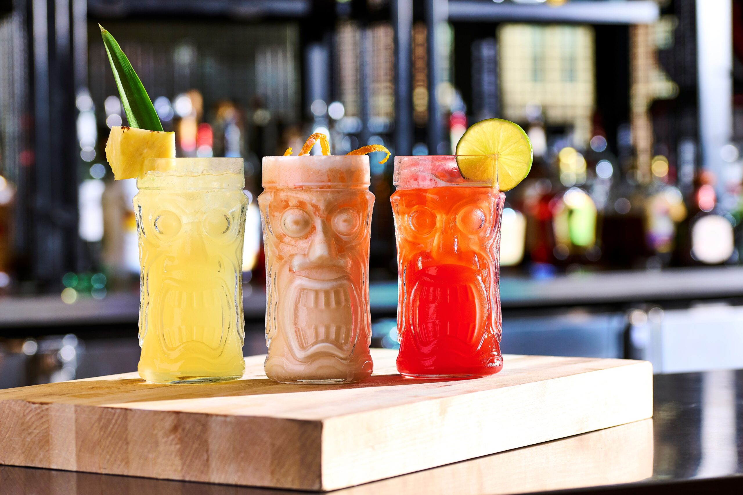 Three colorful tiki drinks sit on a wooden cutting board, garnished with pineapple, an orange twist, and lime. A vibrant bar backdrop enhances the festive vibe.