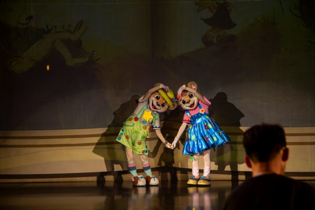 Two cheerful costumed characters resembling anthropomorphic rabbits perform on stage, engaging with an audience member seated nearby.
