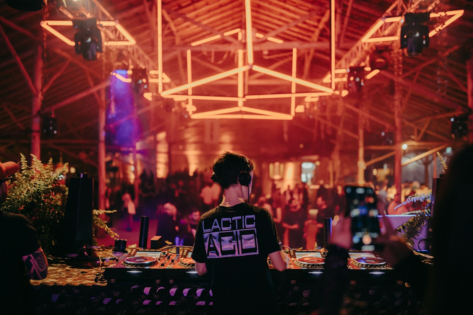 A DJ performs at a lively event, illuminated by vibrant neon lights, while an engaged crowd dances in the background.