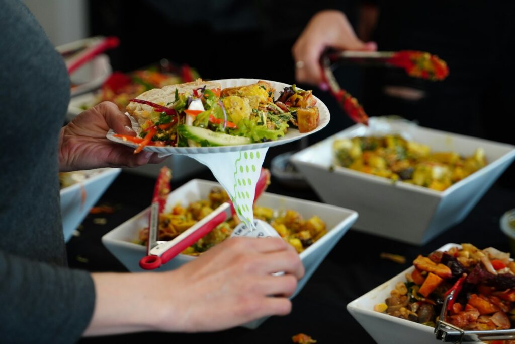 A hand holds a plate filled with colorful salad and garnishes while another hand serves food from a nearby bowl at a buffet.