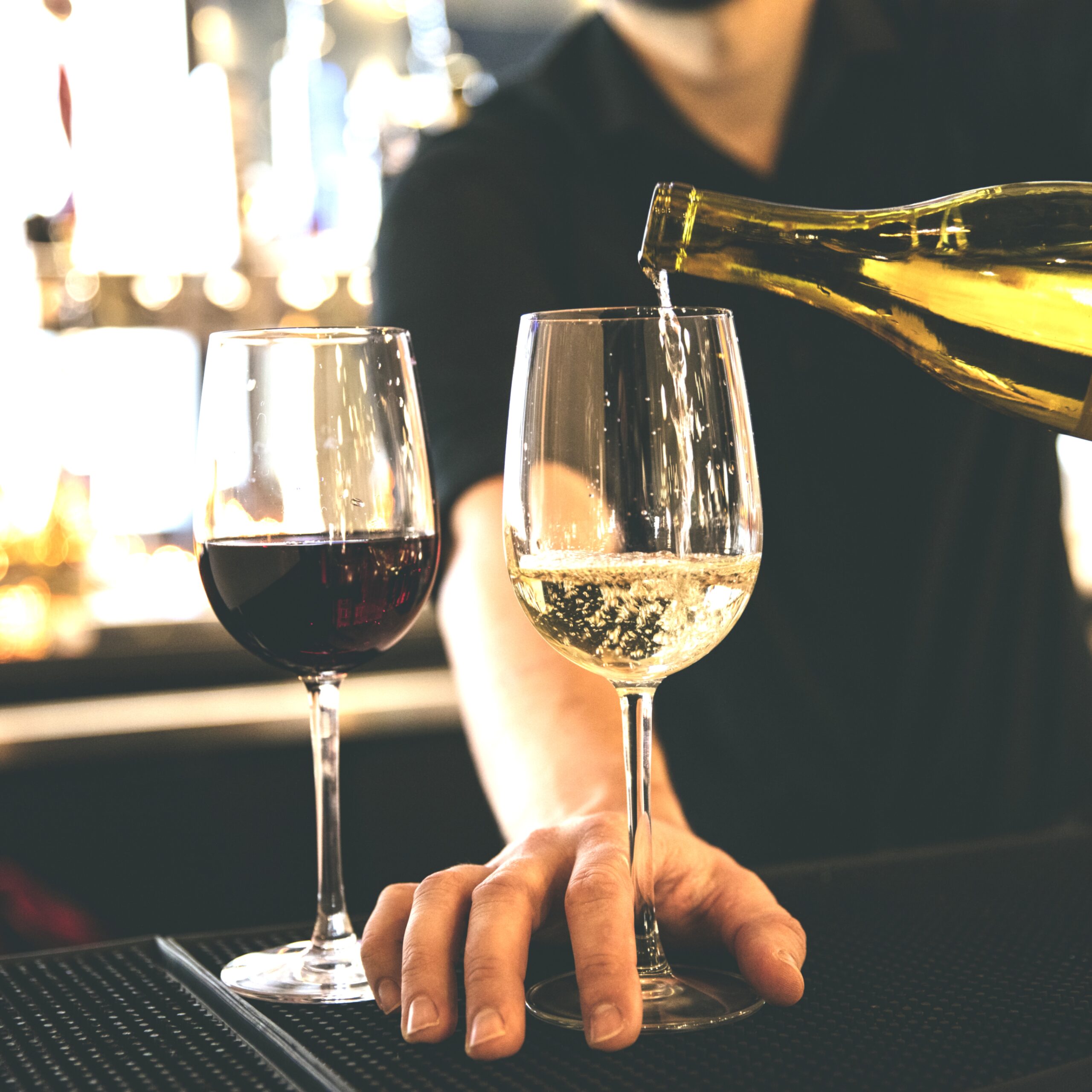 A bartender pours sparkling white wine into a glass beside a red wine glass, showcasing a lively bar atmosphere.