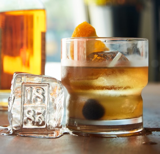 Craft cocktail served in a clear glass, featuring a large ice cube with "1883" embossed on it, garnished with an orange twist and cherry.