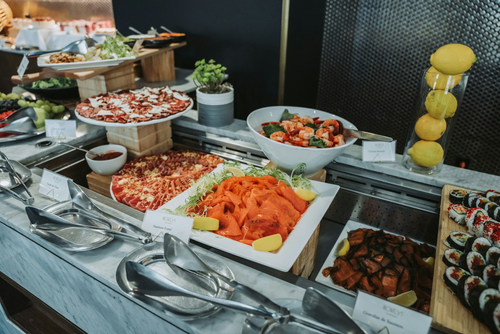 A vibrant buffet display featuring a variety of dishes: smoked salmon, shrimp salad, sushi, and charcuterie on a marble countertop.