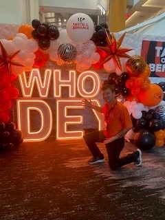 A person kneels beside illuminated "WHO DEY" letters decorated with black, orange, and white balloons, celebrating a Bengals-themed event.